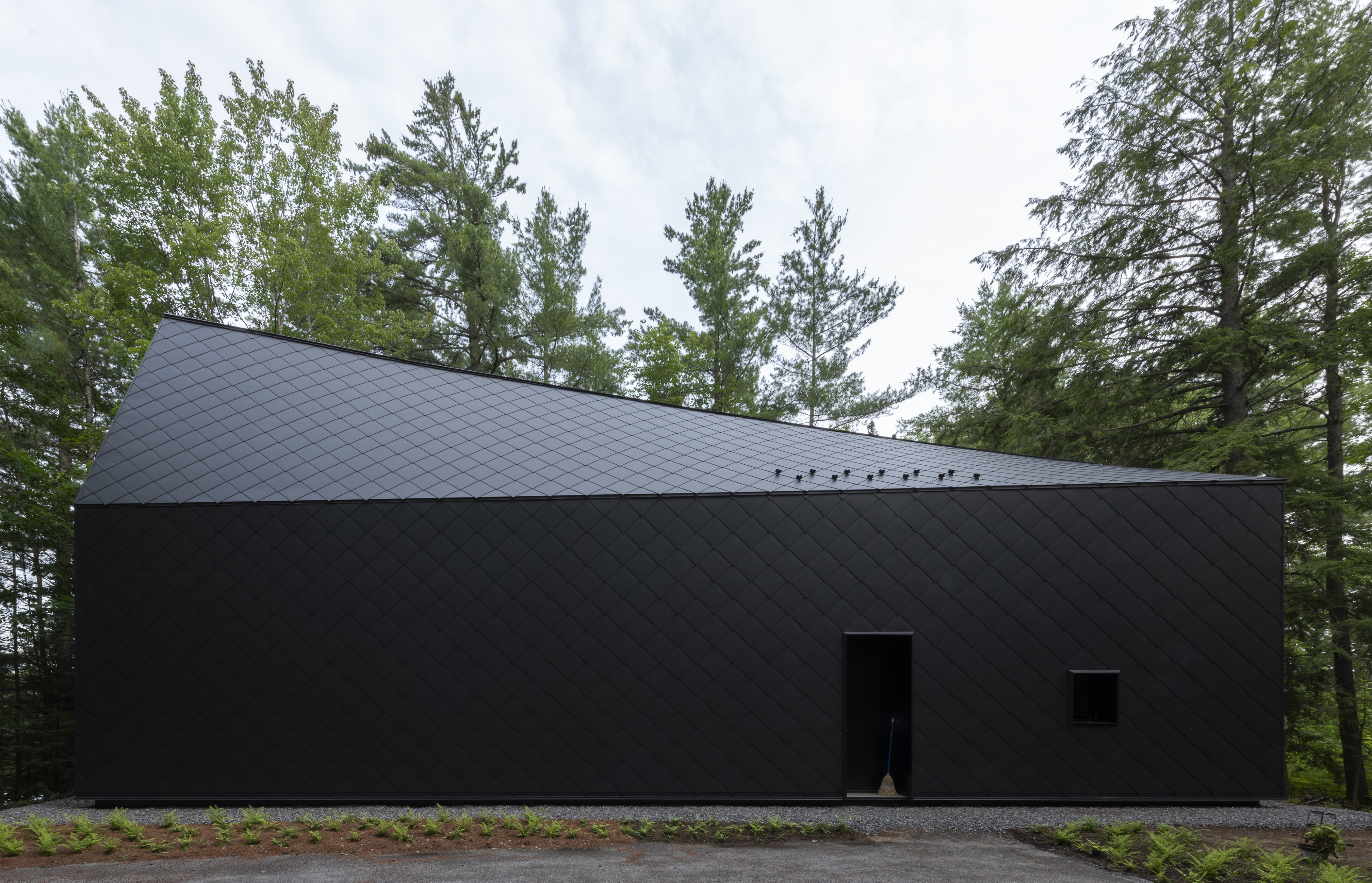 A dark, geometric building with a sloped roof and diamond-patterned exterior panels, set against a backdrop of tall trees and overcast sky