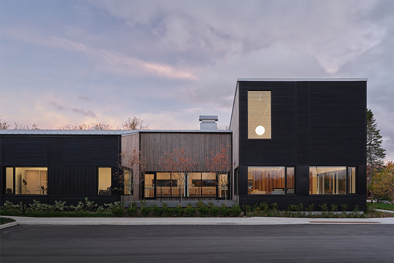 A modern, low-rise building with dark exterior siding and large windows, shown at dusk