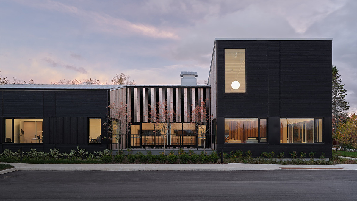 A modern, low-rise building with dark exterior siding and large windows, shown at dusk