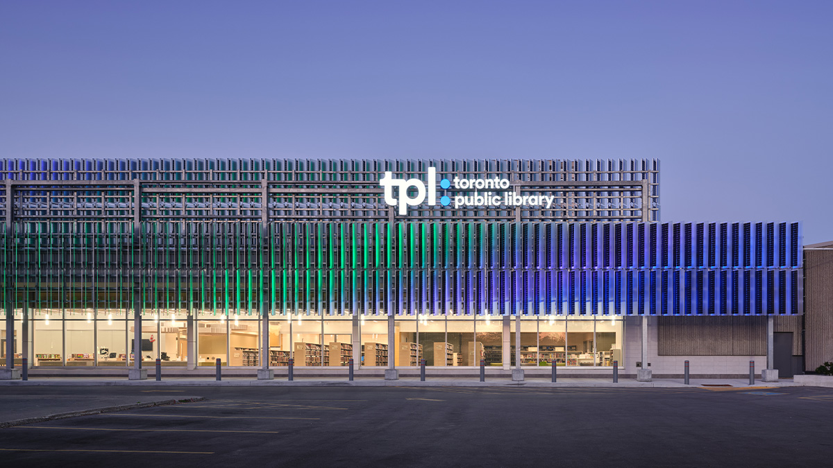 A public library building with a horizontal façade made of vertical metal fins in gradients of green, blue, and purple, large windows on the ground floor reveal bookshelves and interior lighting