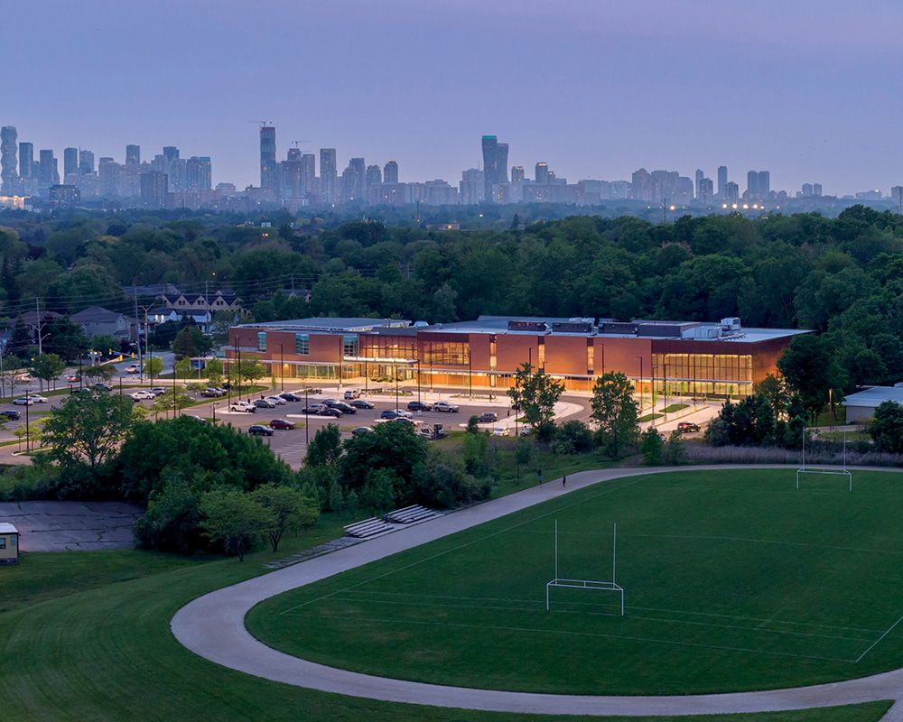 A large modern building sits beside a sports field with the city skyline in the distance