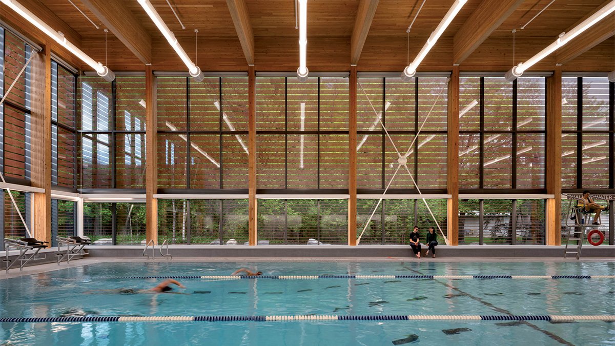 An indoor swimming pool with lane markings, surrounded by tall glass windows and exposed wooden beams