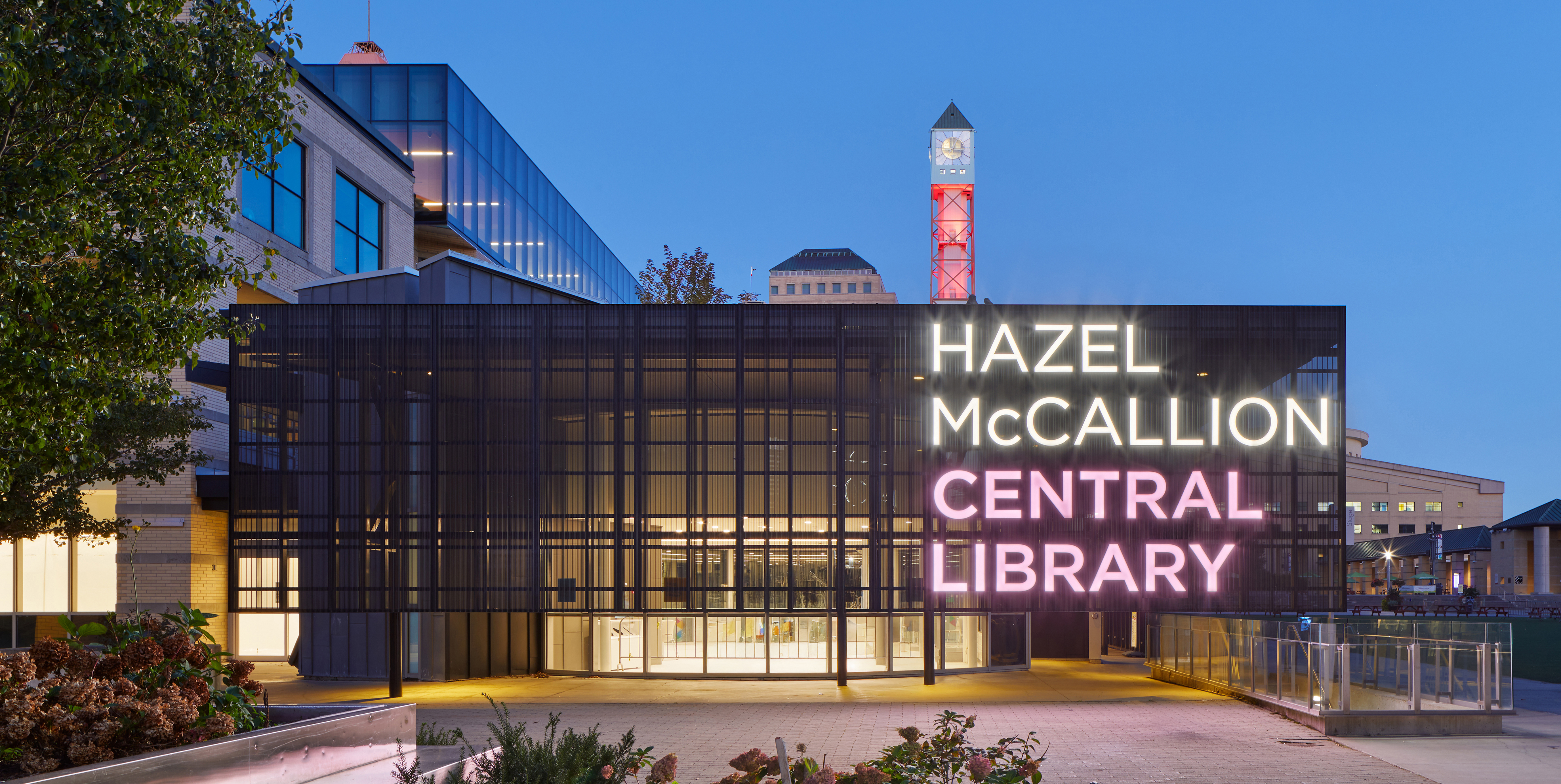 An illuminated modern library entrance at dusk featuring a dark metal façade with large glass panels and bright neon signage reading “Hazel McCallion Central Library,” with a clock tower and surrounding buildings visible in the background