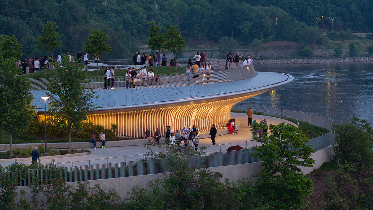 A curved, elevated waterfront overlook with a glowing underside, surrounded by trees and walking paths
