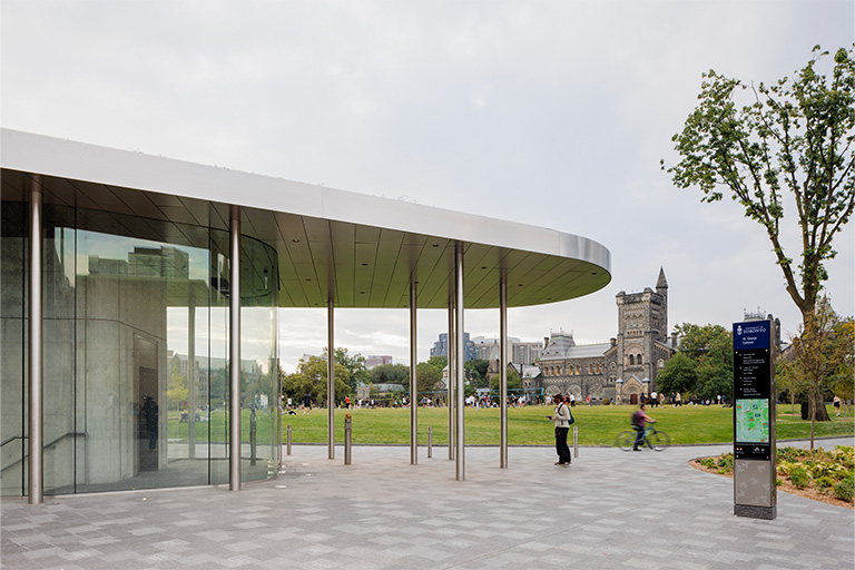 A paved public plaza with people walking near a sleek glass pavilion, with trees showing fall colors