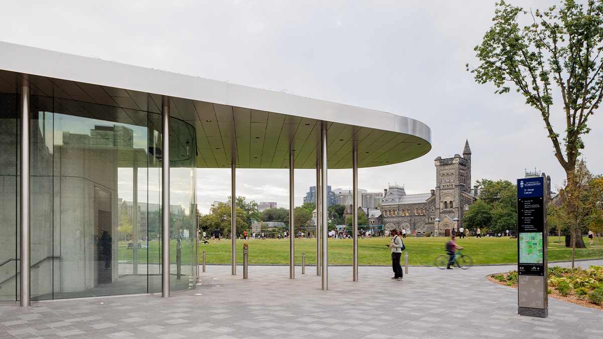 A paved public plaza with people walking near a sleek glass pavilion, with trees showing fall colors