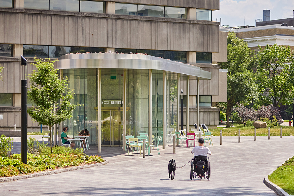A modern glass‑front building with outdoor seating and a person in a wheelchair moving along the path