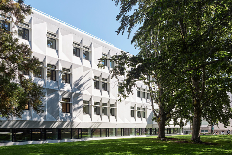 A white institutional building with geometric windows surrounded by a lawn and trees