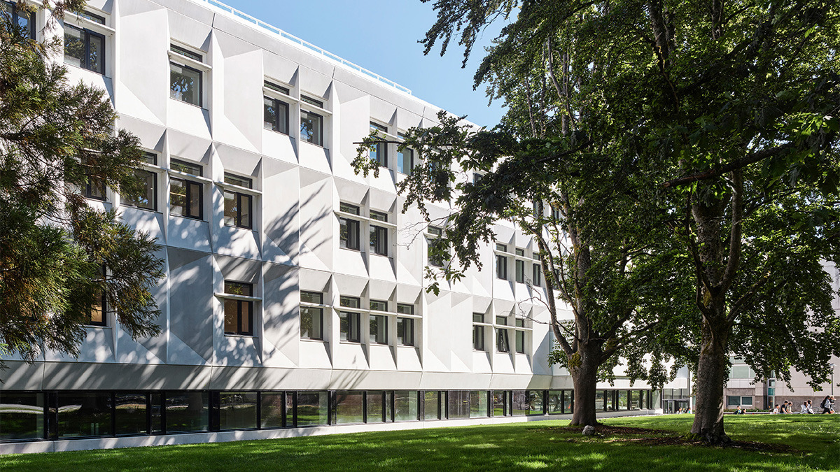 A white institutional building with geometric windows surrounded by a lawn and trees
