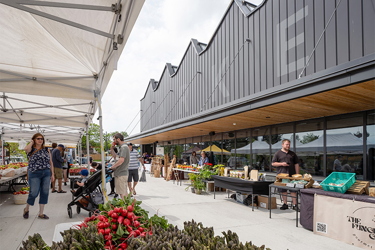 An outdoor farmers’ market with vendor tents set up along a building with a sawtooth roofline