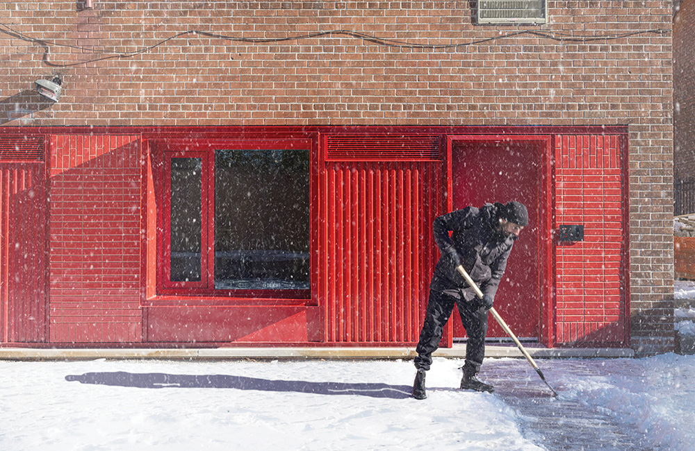 A man shovels snow in front of the corner of the building with a red wall and door