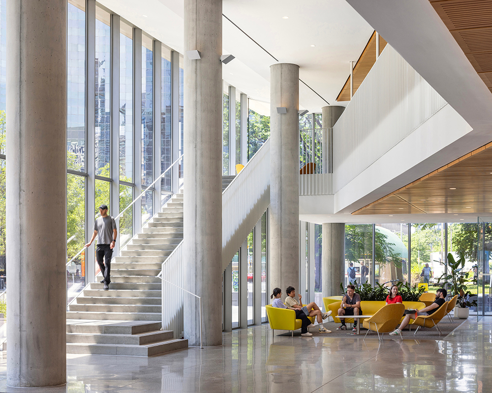 A bright, open atrium with tall concrete columns, floor‑to‑ceiling windows, and colorful lounge seating where people are gathered