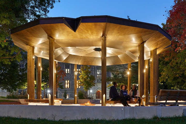 An illuminated pavilion with a wide, faceted roof supported by tall columns, shown at dusk in a park setting