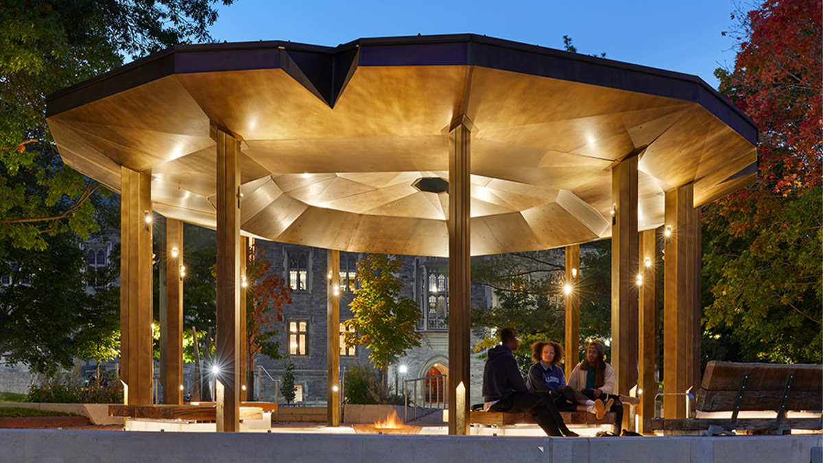 An illuminated pavilion with a wide, faceted roof supported by tall columns, shown at dusk in a park setting
