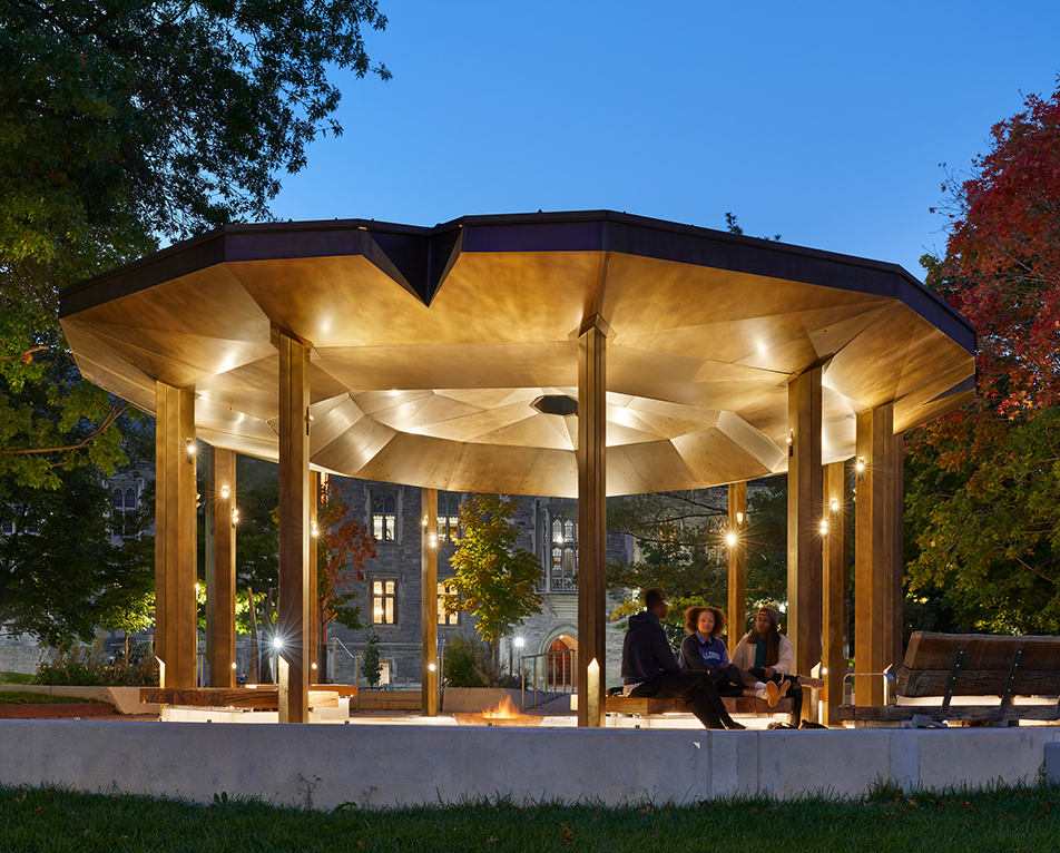 An illuminated pavilion with a wide, faceted roof supported by tall columns, shown at dusk in a park setting
