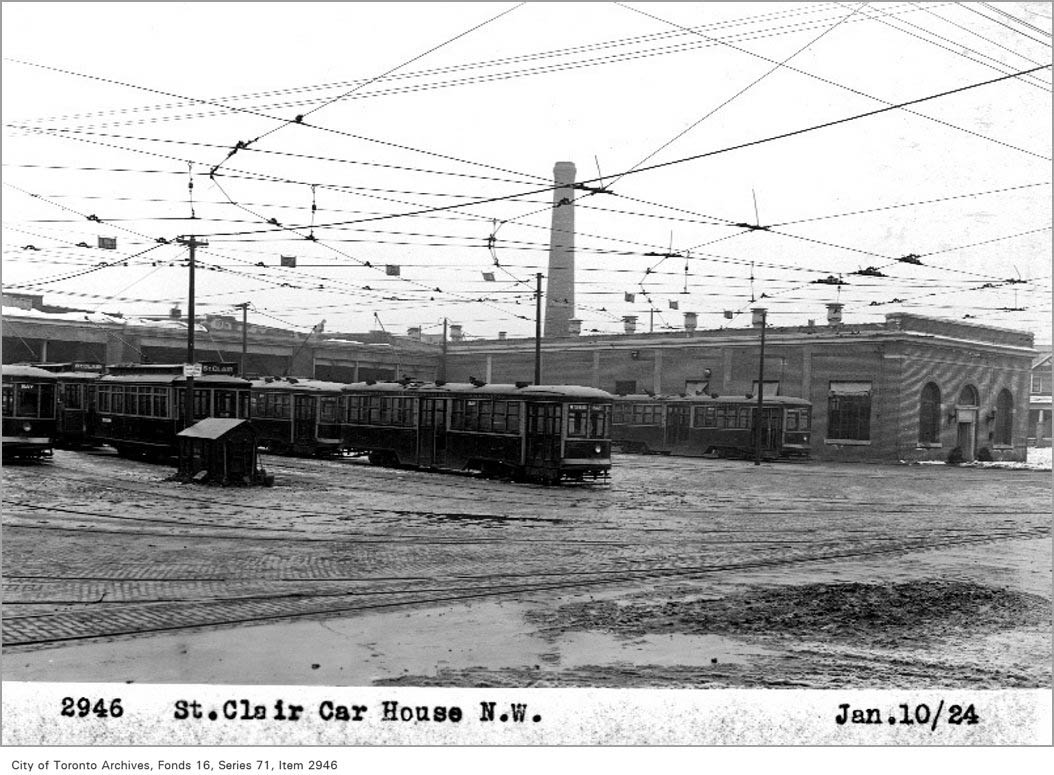 In black and white, three streetcars are stationed in a muddy yard in front of the five car-barns.