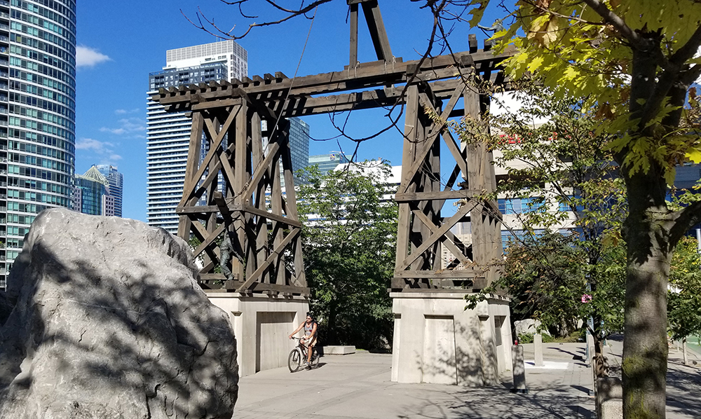 Two wood frame piers on concrete bases are spanned by a railway track. A cyclist passes in between the piers