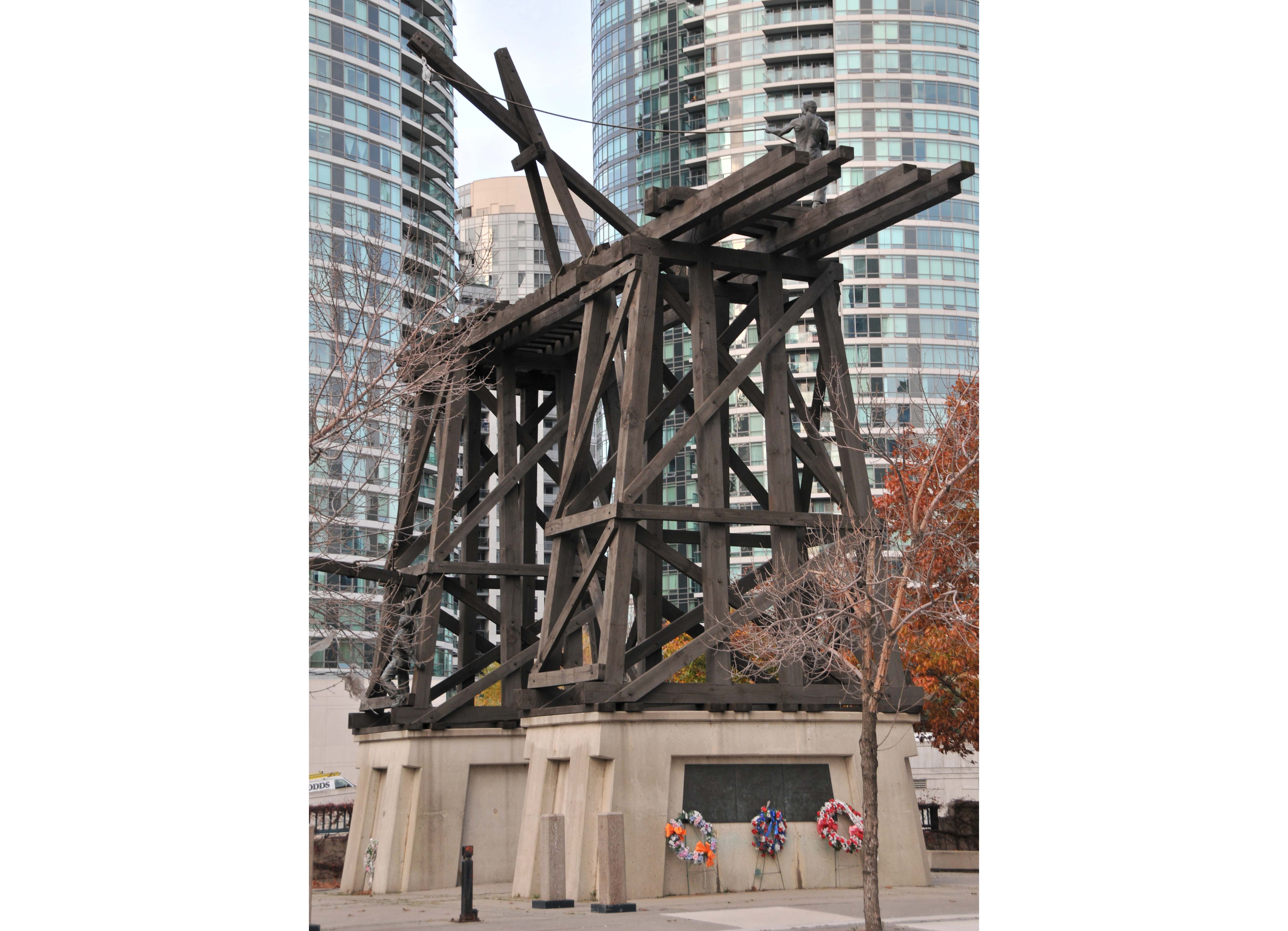 Two wood-framed supporting members spanned by a railway track are being constructed by two copper figures. Three flower wreaths hang on the concrete bases.