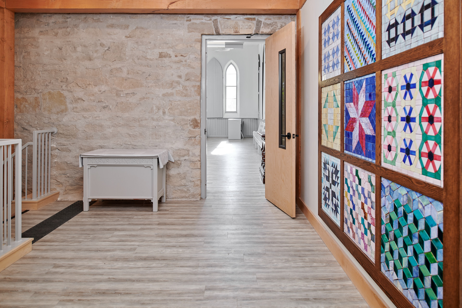 A one-point perspective in a brightly lit interior space with light wood flooring. Another brightly lit space is visible through an open wooden door in a stone wall and, on the right, a large grid of nine quilts is framed on the wall.
