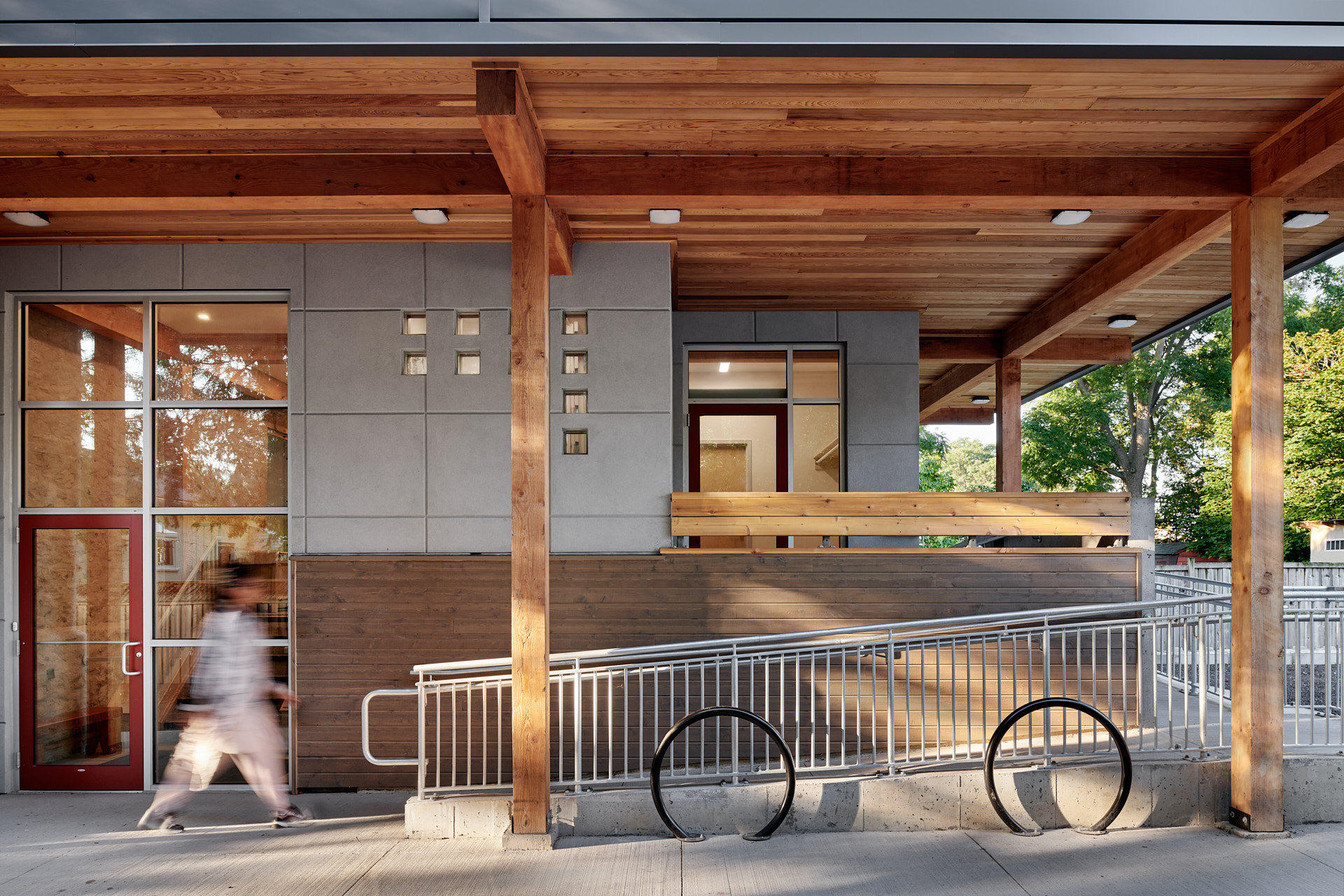 A blurred person walks up an accessibility ramp along the side of the building underneath a large roof overhang supported by heavy timber framing. In the concrete facade of the building is a glazed door, windows, and rows of glass blocks arranged in a pattern.