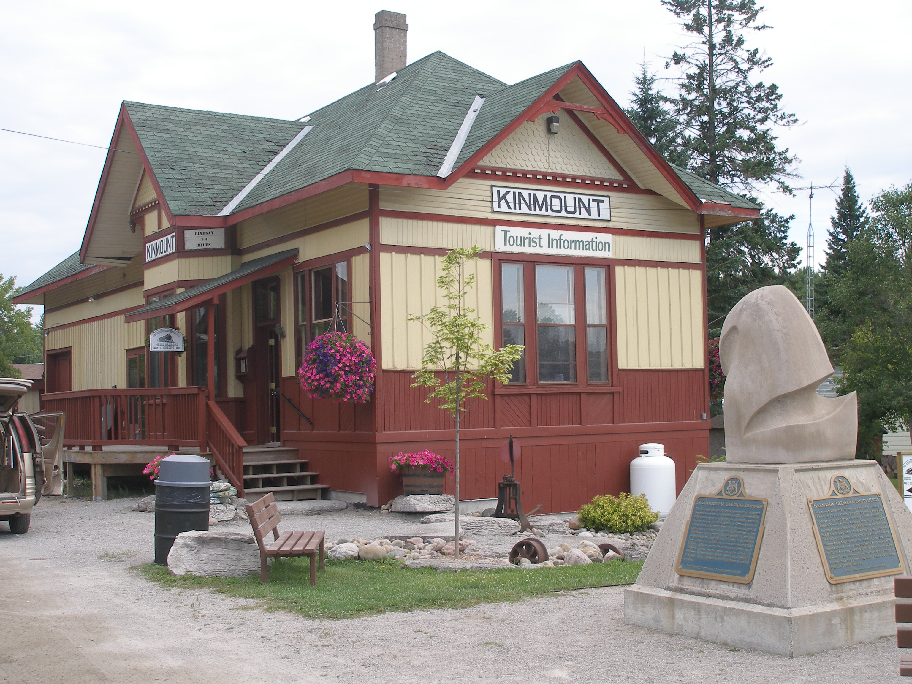 Small railway station building with hipped roof and wooden side panelling. A small abstract sculpture on a plinth stands in the foreground.