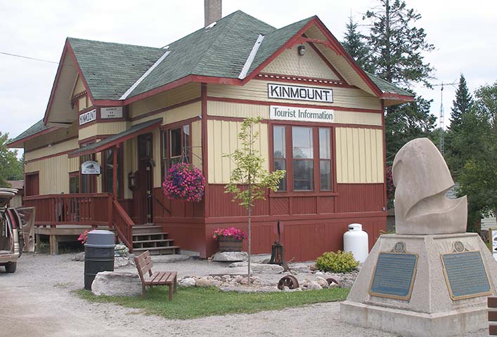 Small railway station building with hipped roof and wooden side panelling. A small abstract sculpture on a plinth stands in the foreground.