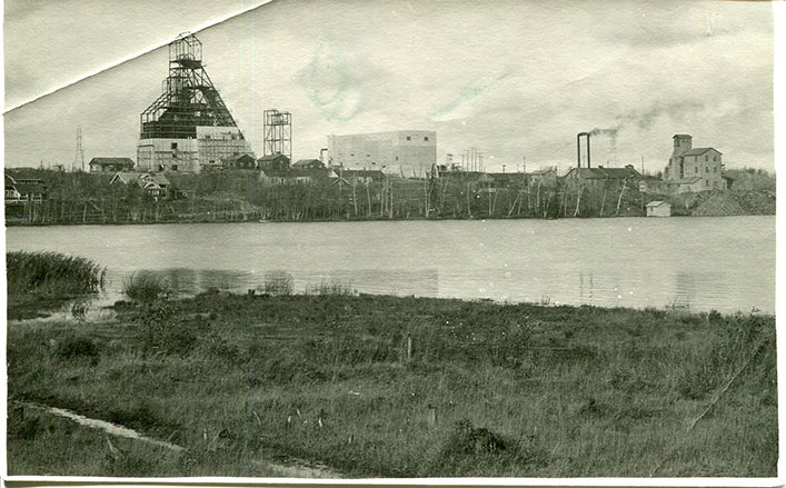 Mining site with homes, smokestacks and other structures at edge of a lake, with large headframe under construction. 