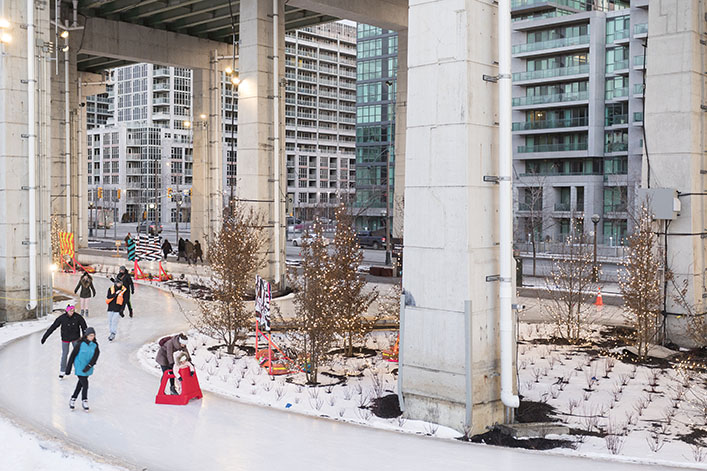 People skating along a path located under an elevated highway, with condominiums in the background. 