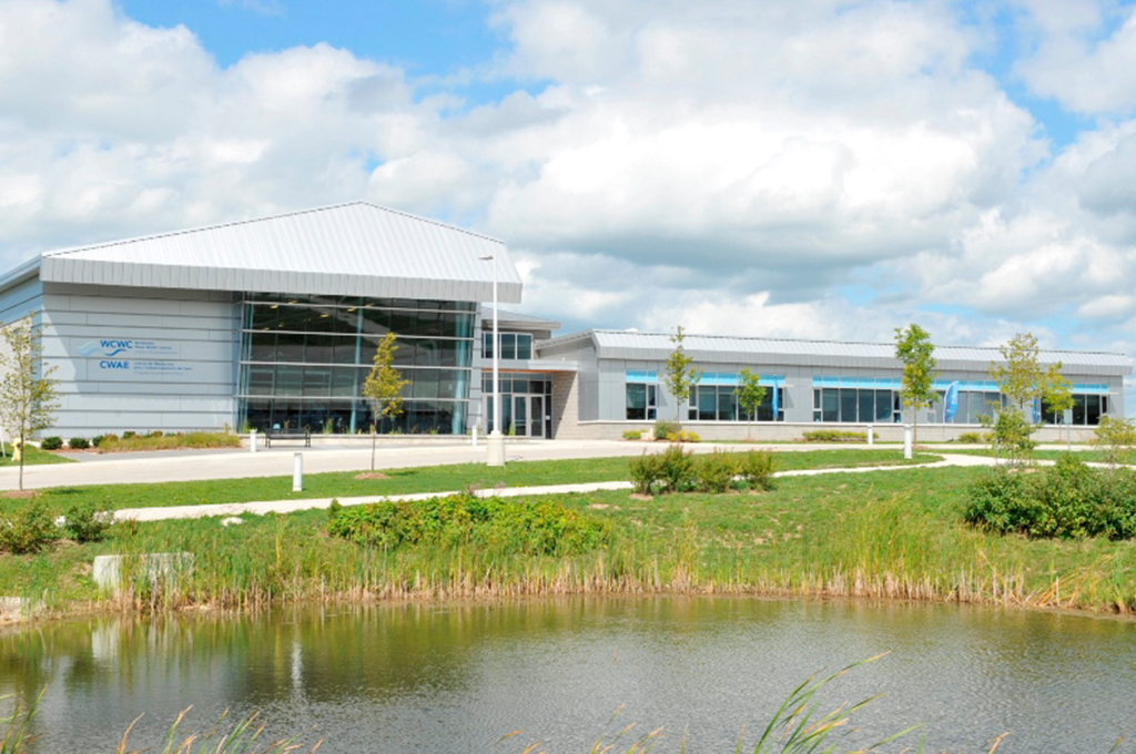 Exterior view of Walkerton Clean Water Centre’s building, a low one storey contemporary building with a large windows situated in front of a stormwater retention pond and young vegetation