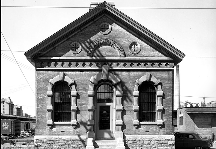 Front elevation of a neoclassical building featuring rounded arch windows and door, a brick facade and rusticated stone at the bottom. In the centre over the door is a stone carving that reads City Registry Office. 