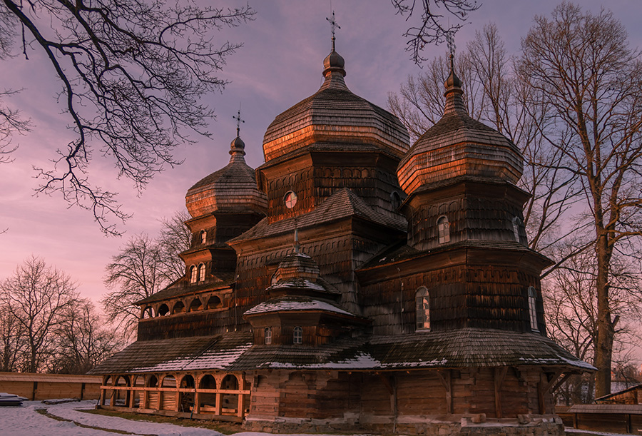 Wooden church with ground floor arcade, overlapping roofs, and three upward projecting onion domes topped with crosses.