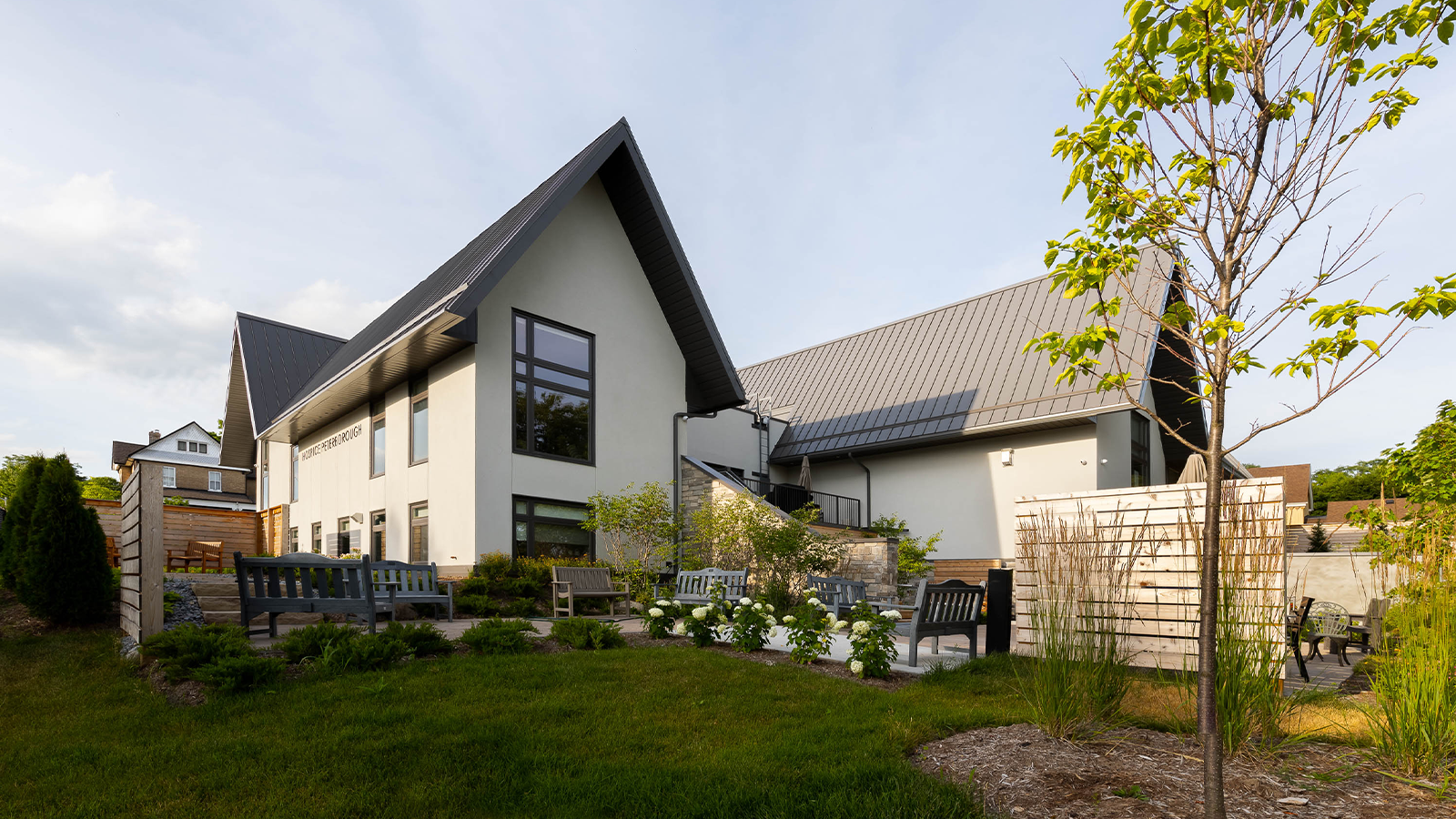 Medium-sized white stucco building with tall, steep gable roofs made of dark grey metal. In front, a garden with seating and some planting.