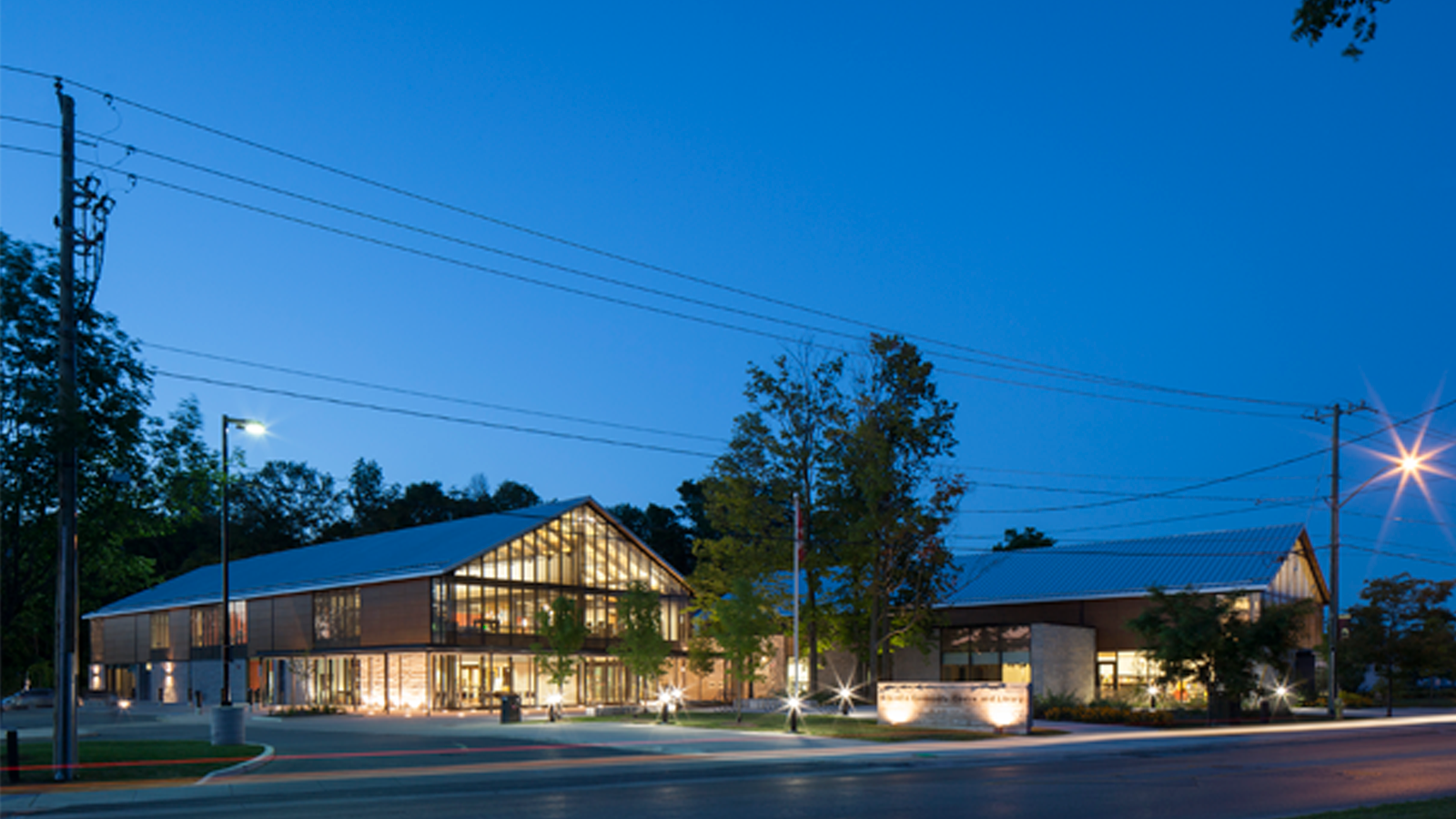 Two large, low, interconnected buildings with metal gable roofs and glass and stone walls. The buildings glow with warm light against an evening sky highlighting the sillouhette of trees around them.