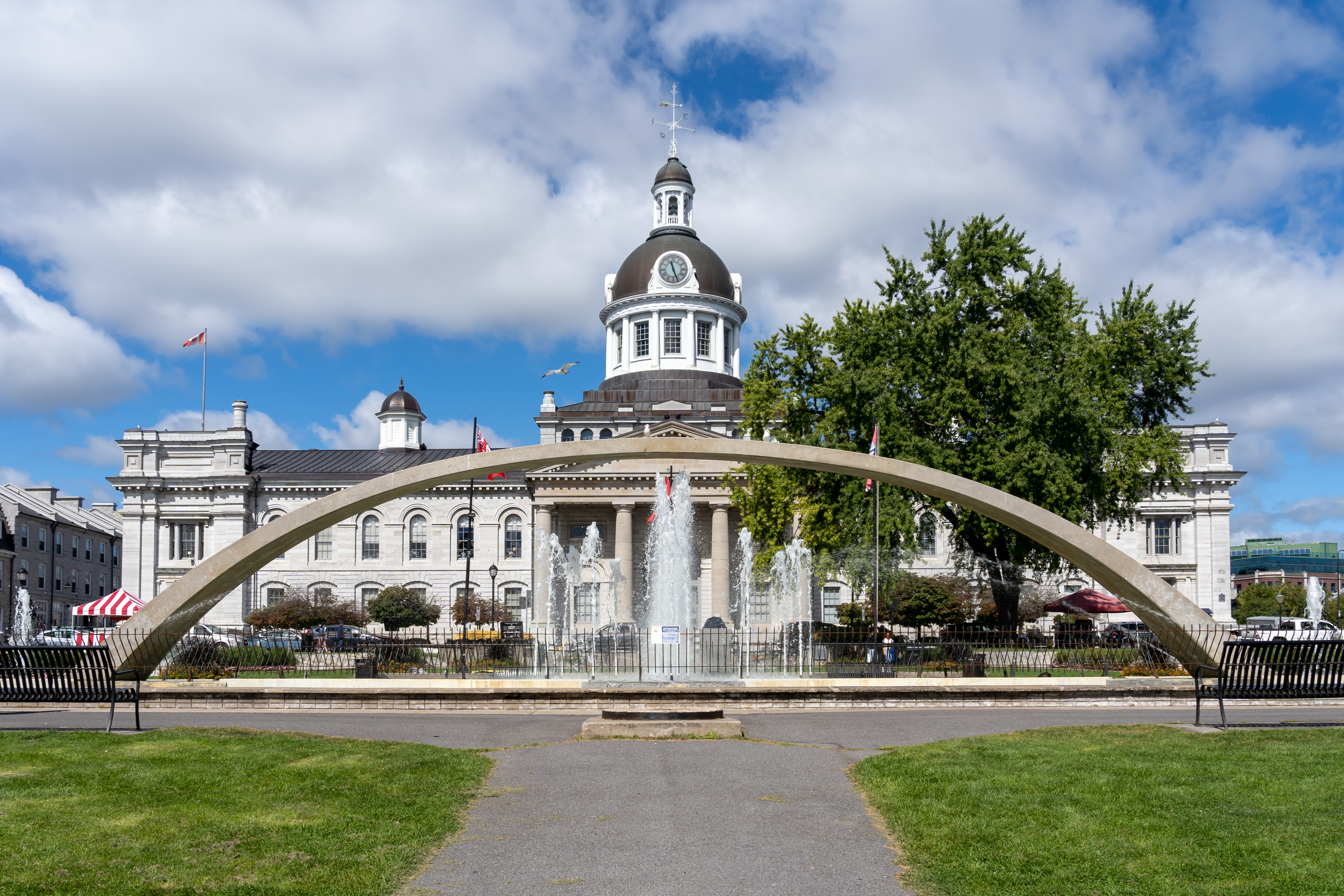 A large limestone building with a prominent central cupola and dome containing a clock and weather vane. In front, a large, contemporary arched concrete water fountain. 