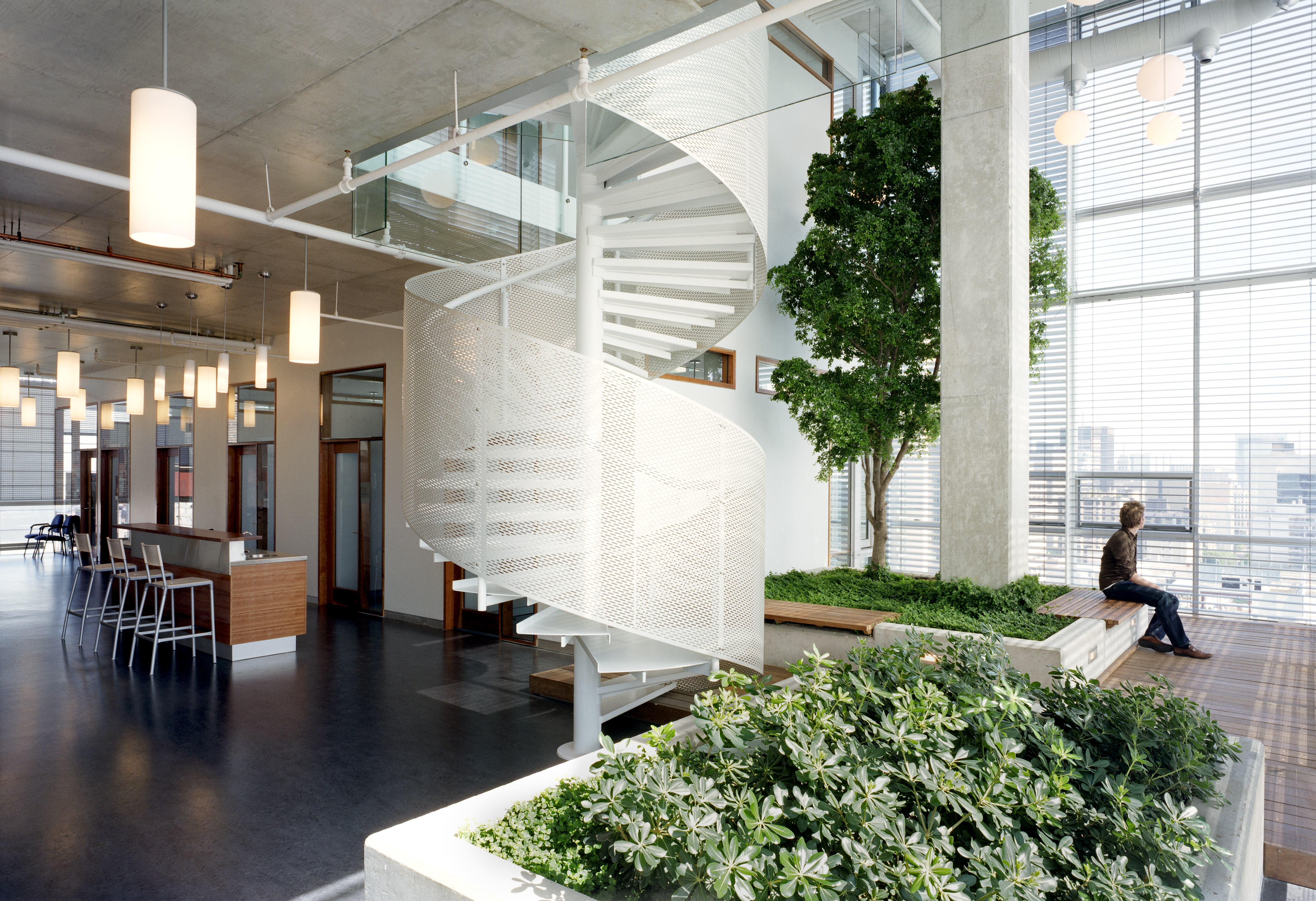 A bright, double-height indoor garden with a white metal spiral staircase, plants, a tree, and wood benches that are integrated into the sides of concrete planters.										