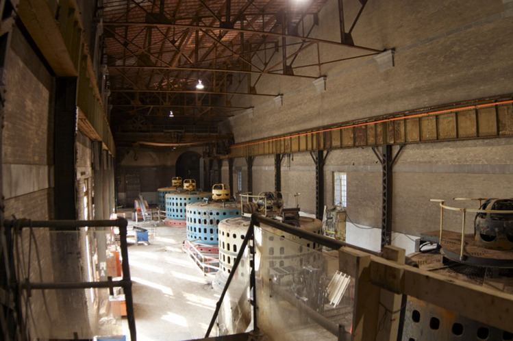 A tall open warehouse space with four round generators in a row, one grey and three blue. 