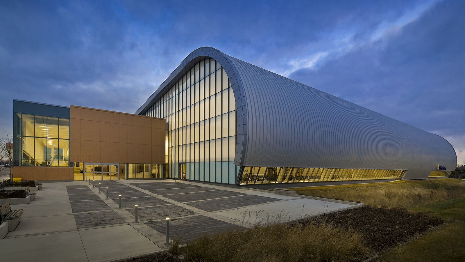 Large, curved, metal-clad structure with curtain wall windows adjoining a rectangular metal and glass building.